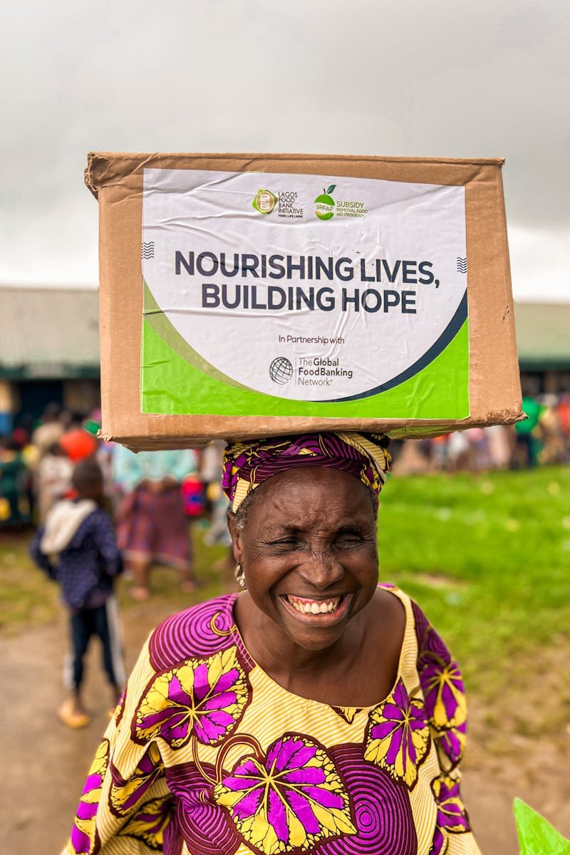 Elderly woman joyfully holding a donation box on her head outdoors, spreading hope.