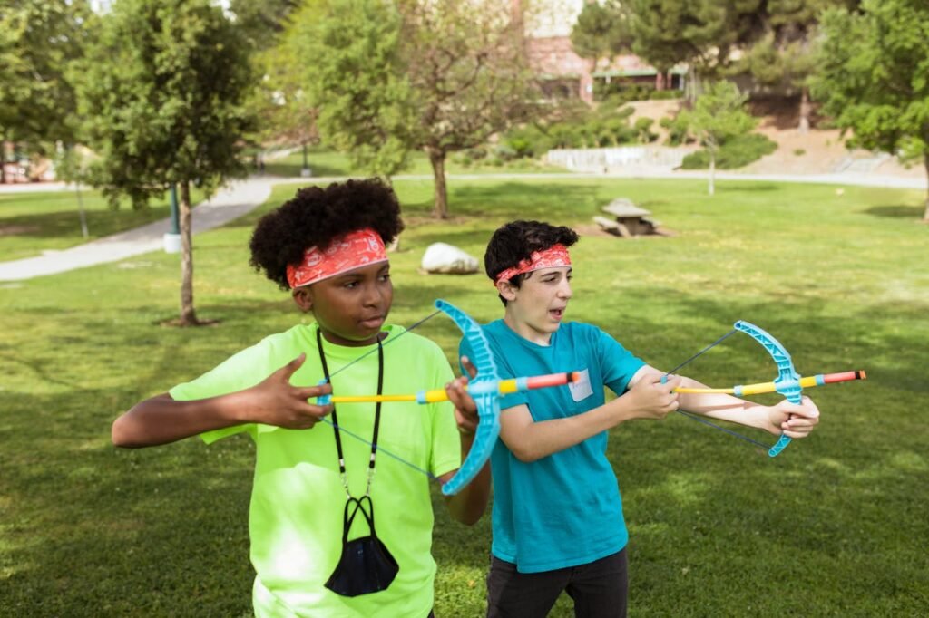 Two children practicing archery outdoors, having fun and learning teamwork at summer camp.